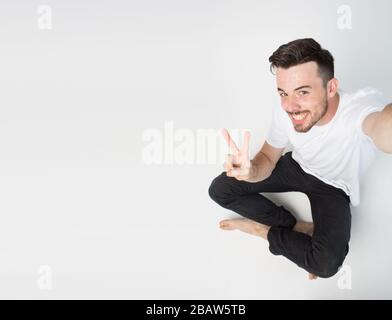 Young man isolated over background. Guy sit on floor and take selfie. Hold camera with one hand and show peace symbol. Emotional happy guy on picture. Stock Photo
