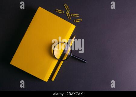 Yellow pencil, yellow notebook glasses, pins, clip paper and a magnifying glass on on black background Stock Photo