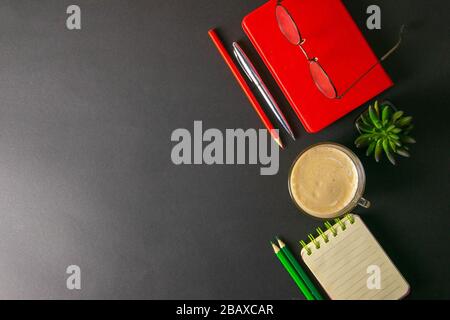 The red notebook stands on a black background, on the notebook there are glasses and next to the notebook, a flower, coffe, pen and a red pencil Stock Photo