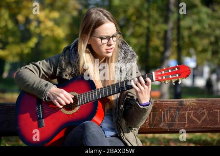 Young, cute girl in jeans sitting on a bench in a park and playing red acoustic guitar Stock Photo