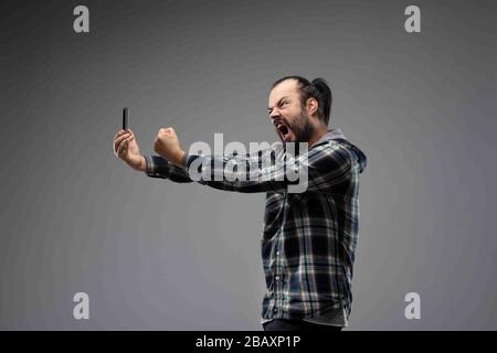 Bearded man in checked shirt menacingly and aggressively screaming at the phone and showing clenched fist. Arguing online concept portrait, viewed fro Stock Photo