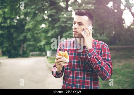 Handsome young man eating sandwich autdoor. He is holding a phone Stock Photo