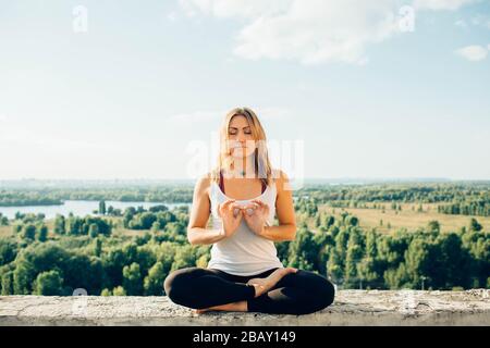 Young woman practices yoga outside. Calm smiling girl sits on parapet in lotus position with close eyes. Her fingers fold into sign of infinity Stock Photo