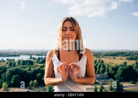 Young woman practices yoga outside. Calm smiling blonde girl with close eyes. Her fingers fold into sign of infinity. Portrait. Nature trees river and Stock Photo