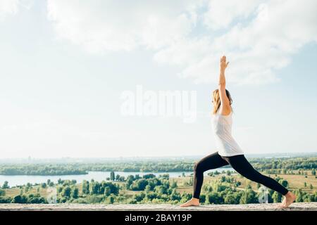 Young woman practices yoga outside. Blonde girl standing in Warrior I posture Virabhadrasana 1 , full length on parapet on beautiful view. Trees river Stock Photo