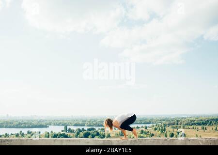 Young woman practices yoga outside. Blonde girl do crane pose or bakasana on parapet with beautiful view. She dressed in black leggings and white T Stock Photo