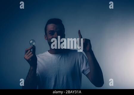 Young brunette man with beard dressed in white T-shirt. He smiles broadly. Guy holding light bulb in right hand another points finger up. Isolated Stock Photo