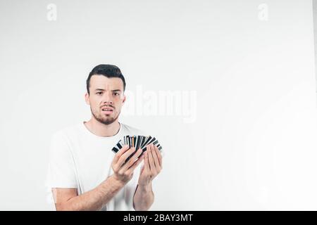 Young brunette man with beard dressed in white T-shirt. Guy holds in hands fan of credit cards. His face unpleasantly surprised. Isolated over light Stock Photo