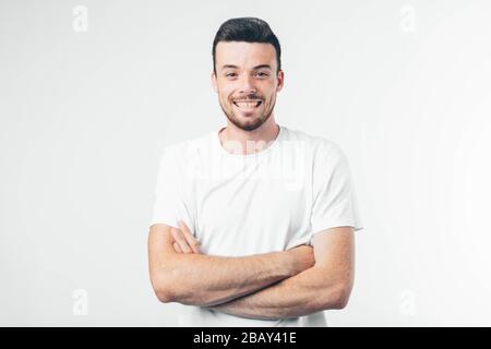 Young brunette man with beard dressed in white T-shirt. Guy confident and wide smiling. Arms crossed at chest level. Isolated over light background. Stock Photo