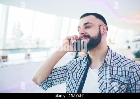 Side view of young bearded man,dressed in gray cardigan,sitting at round wooden table in cafe with modern interior and is holding smartphone Stock Photo