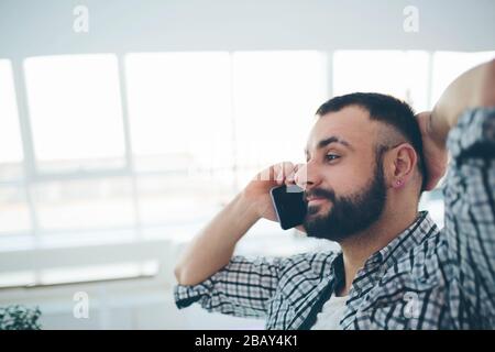 Side view of young bearded man,dressed in gray cardigan,sitting at round wooden table in cafe with modern interior and is holding smartphone Stock Photo