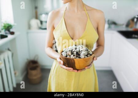Woman in yellow dress holding in hands wooden bowl with pile of organic quail eggs. Easter cooking concept. Kitchen on background. Stock Photo
