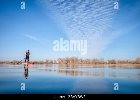 senior male paddler in a wetsuit is paddling a stand up paddleboard on a lake in Colorado, winter or early spring scenery, low angle action camera vie Stock Photo