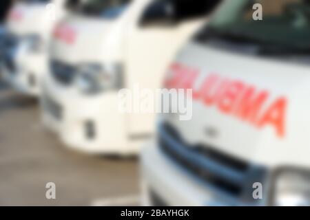 A blurred front part of ambulances standing in parking lot of hospital. A defocused row cars of rescuers with red text ambulance on hood. Stock Photo