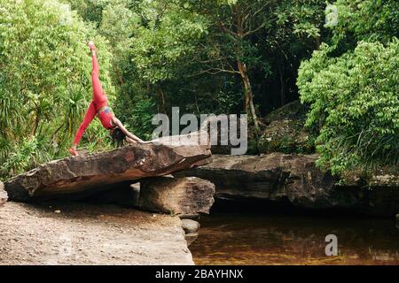 Flexible slim yuong woman bending forward and raising leg when practicing yoga in forest Stock Photo