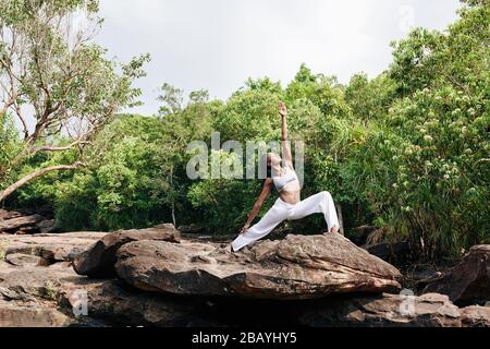 Fit Black woman practicing yoga on big rock and doing warrior pose Stock Photo