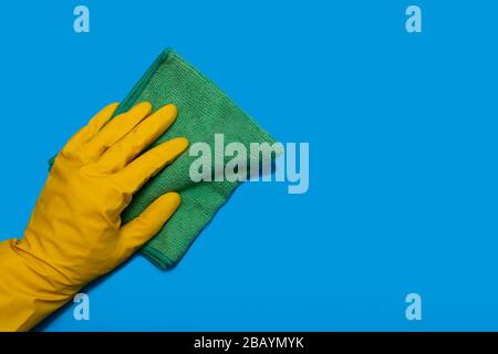 Hand in yellow glove with green rag of microfiber wipes on blue background. Copy space. Horizontal Stock Photo