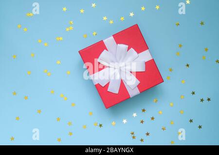 red gift box with white bow on color blue background with star shaped confetti. Festive greeting card. Holiday concept Stock Photo