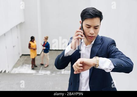 Young Asian businessman talking on phone and checking time on his wrist watch Stock Photo