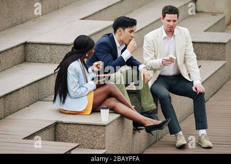 Business colleagues sitting on steps outdoors, eating lunch and discussing news and work Stock Photo