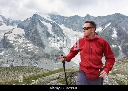 Portrait of a man wearing a red jacket and a backpack, having a walk with hiking poles in the mountains situated in Swiss Alps, rocky ridge is on background