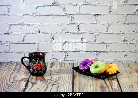 A rectangular plate with five multi-colored donuts and a black ceramic coffee mug stand on a wooden table. Close-up. Stock Photo