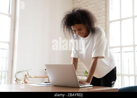 Happy mixed-race businesswoman leaning over office desk chatting online Stock Photo
