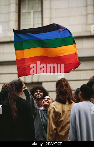 Smiling woman waving gay flag with group of people standing around. People participating in Gay march in the city. Stock Photo