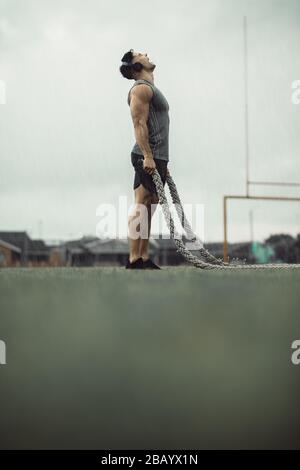 Man taking rest from battle rope workout and screaming. Muscular man doing fitness workout outdoors in rain with battle ropes on a field Stock Photo