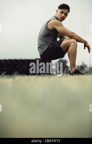 Young man sitting on a big tire outdoors after a workout. Muscular man taking a break from cross training outdoors. Stock Photo