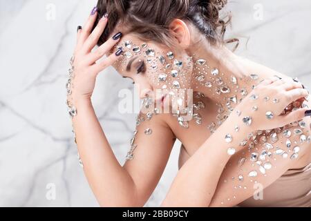 Unusual makeup and face art with sparkling rhinestones on the skin. A girl with precious crystals on her face, a fashion image in light soft colors. Stock Photo