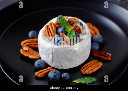 Gourmet raw soft alemar cheese with blueberries, pecans and fresh mint on top on a black plate on a dark concrete table, close-up Stock Photo