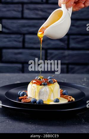 Woman hand pouring dripping honey on top of baked soft brie cheese with pecan, blueberries served on a black plate on a concrete table in front of a b Stock Photo