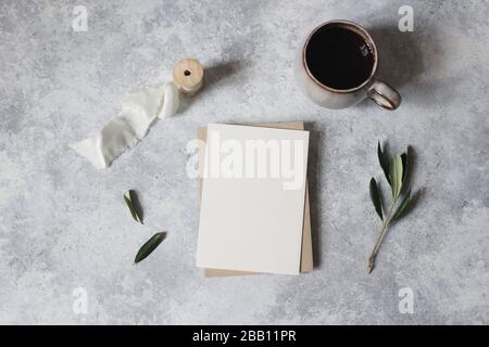 Moody feminine wedding stationery mock-up scene. Blank greeting card, cup of coffee and green olive leaves, branches. Grunge table background. Flat la Stock Photo