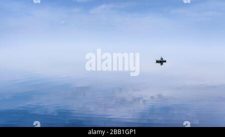 Mirror Surface Lake with a Lone fisherman on an inflatable boat and mice blue sky on background Stock Photo