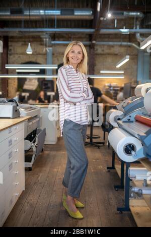 Blonde woman standing near paper roll machine with folded arms, smiling Stock Photo