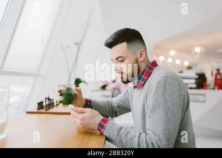 Young bearded man in cafe. He dressed in gray pullover and plaid shirt. Man holds tea and looks at smartphone . Chessboard flower and blurred interior Stock Photo