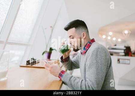 Young bearded man in cafe. He dressed in gray pullover and plaid shirt. Man is drinking tea and using smartphone. Chessboard, flower and blurred Stock Photo