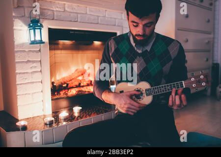 Picture of young man sitting at fireplace and playing ukulele. Alone in room inside practicing. Singing songs and playing melodies. Stock Photo