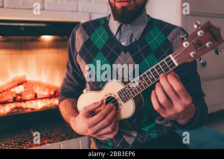 Close up and cut view of young bearded man playing ukulele in room at fireplace. Practicing and having fun alone. Young musician in room. Stock Photo