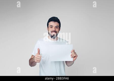 Young man isolated over background. Positive cheerful guy hold white blank page and hold big thumb up. Young hipster in shirt alone in studio. Stock Photo