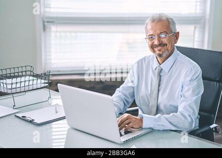 Business portrait of an elderly man. An aged man working on a laptop, looking at the camera and smiling. Work on removal, freelance. Stock Photo
