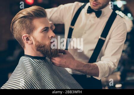 Cropped photo of a classy dressed barber shop stylist working for a perfect look of a red bearded guy in a cape. His hairdo looks stunning! Stock Photo