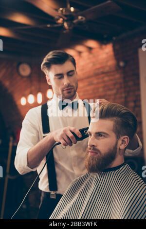 Beard styling for a handsome young guy at the barber shop. Hairdresser is attractive and wearing classic outfit, he is focused and serious Stock Photo