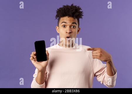 Close-up portrait of excited, cheerful young hispanic male geek talking about his device, new gadget or application, holding mobile phone, pointing at Stock Photo