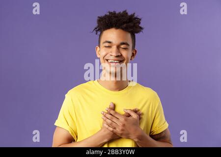 Close-up portrait of happy, upbeat young dreamy guy, remember sweet memories, hold hands on heart, smiling touched and delighted, close eyes grinning