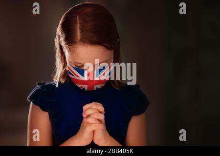 Pray for UK. Child in face mask praying for Great Britain. Little British girl in hospital chapel or church during coronavirus outbreak. Virus pandemi Stock Photo