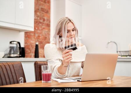 A Single, caucasian female with blonde hair sits at a table and looks at a laptop whilst holding a bank card Stock Photo