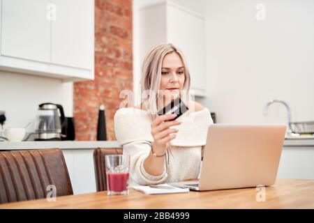 A Single, caucasian female with blonde hair sits at a table and looks at a laptop whilst holding a bank card Stock Photo