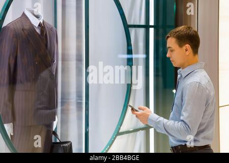 Buyer Caucasian man with a phone stood near a luxurious men's suit in supermarket window Stock Photo